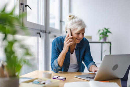 smiling mature businesswoman talking on smartphone and using laptop in officeの写真素材