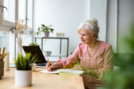 smiling senior woman writing in notebook while working with laptop at homeの写真素材