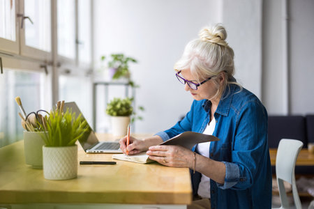 Senior woman writing in notebook while sitting at table in home office.の写真素材