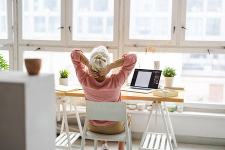 senior woman with back pain sitting at table with laptop at homeの写真素材