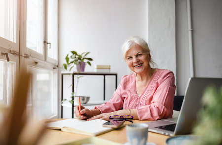 Smiling senior woman sitting at the table and using laptop at homeの写真素材