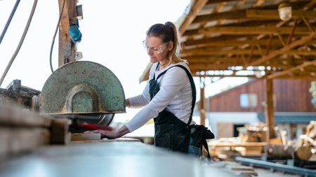 Female carpenter working with circular saw in woodworking workshop. She is wearing black overalls and glasses.の写真素材