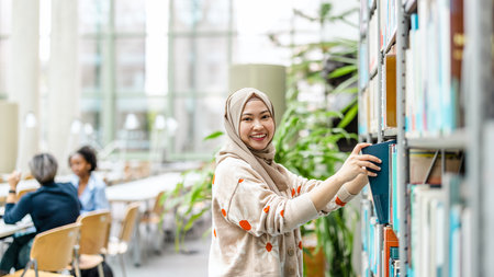 Portrait of young asian muslim woman wearing hijab and smiling while standing in libraryの写真素材