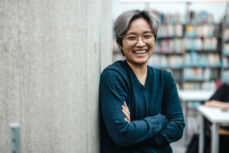 Portrait of happy Asian college student standing with arms crossed in libraryの写真素材