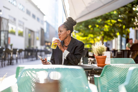 Young businesswoman drinking orange juice and using mobile phone in a street cafeの写真素材