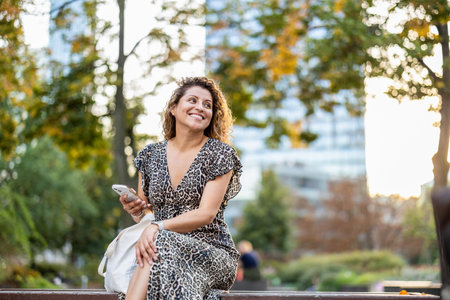 Beautiful young woman sitting on a bench in the park and using mobile phoneの写真素材