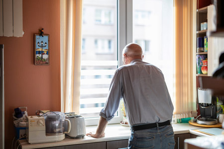 Mature man standing at the kitchen counter in his house and looking out the windowの写真素材