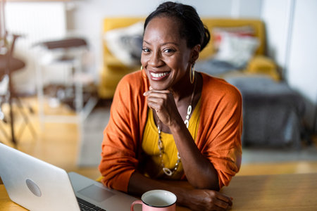 Portrait of smiling woman using laptop at table in living room at homeの写真素材