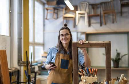 Portrait of young female carpenter standing in workshop and using mobile phoneの写真素材