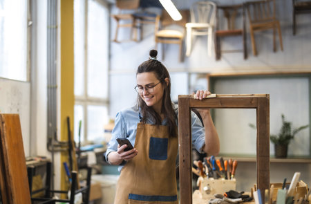 Portrait of young female carpenter using mobile phone in her workshopの写真素材