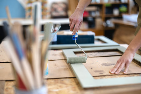 Close-up of female artist using paint roller to paint wooden plank in workshopの写真素材