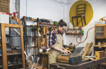 Portrait of female carpenter using laptop in workshop at her workplaceの写真素材