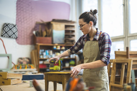 Carpenter working on wood in her workshop. She is wearing apron and glasses.の写真素材