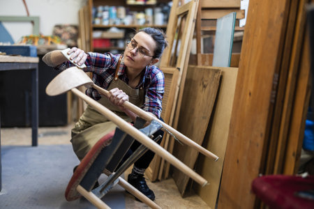 Female craftsman working on a wooden chair in a workshop, wearing glassesの写真素材