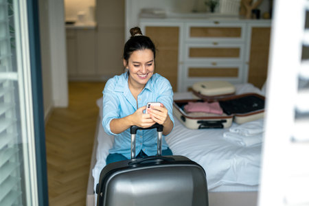 Smiling young woman using mobile phone while sitting on suitcase in hotel roomの写真素材