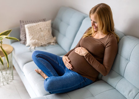 Pregnant woman sitting on sofa and touching her belly at homeの写真素材