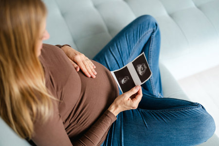 cropped shot of pregnant woman holding ultrasound scan on sofa at homeの写真素材