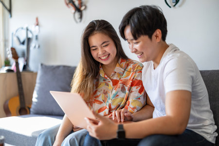 Happy asian couple using digital tablet and smiling while sitting on sofa at homeの写真素材