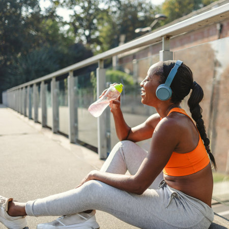 Sporty african american woman drinking water while sitting on bridgeの写真素材