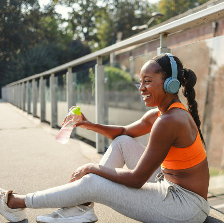 Happy african american sportswoman drinking water while sitting on bridgeの写真素材