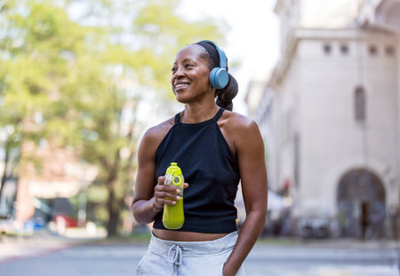 Portrait of happy african american woman listening to music with headphones while holding water bottle in cityの写真素材