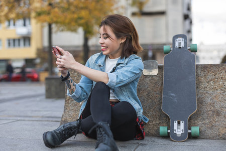 Young woman with skateboard sitting on a pavement and looking at her mobile phoneの写真素材