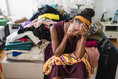 Tired african woman sitting on bed with pile of clothes in bedroomの写真素材