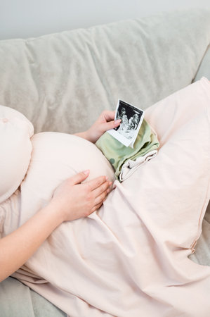 Vertical shot of a young pregnant woman sitting on sofaand checking sonogram.Side view