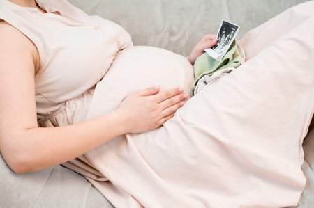 Unrecognizable pregnant woman sitting on sofaand checking sonogram.Side view