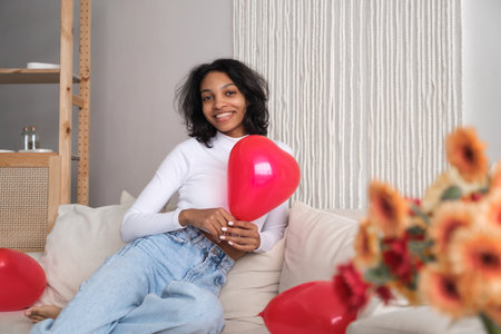 Young african american woman holding red heart shaped balloon sitting on sofa at home. Celebrating valentines, love day, women's day, birthday. Copy space bannerの写真素材