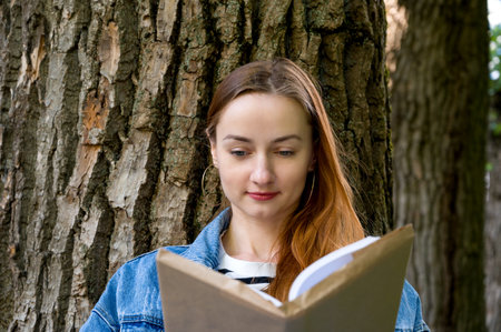 Education or relaxing concept. Young red haired female reading book in the park under the tree