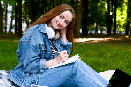 Red haired caucasian woman studying remotely on laptop, writing down information in notebook, outside.Education concept. home workの写真素材