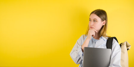 Portrait of thinking young female with laptop with confused puzzled face on yellow isolated...