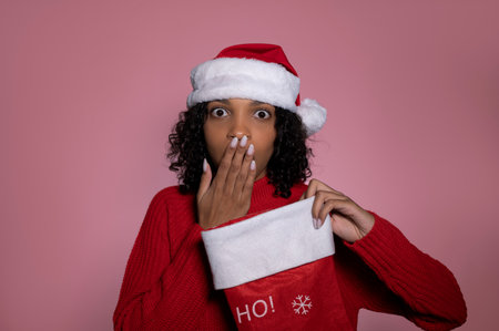Surprised dark skinned santa girl with stocking on pink background.Merry christmas and winter holidays concept.の写真素材
