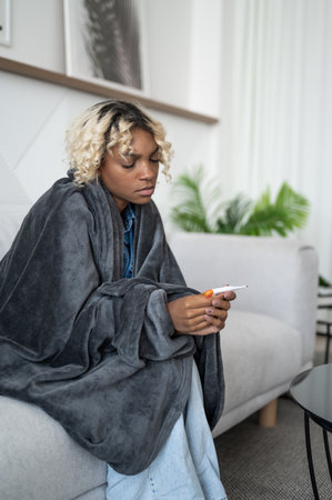 Young ill woman sitting on the couch under the blanket holding thermometer.Flu and cold season