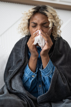 Closeup of Sick young black woman sitting under the blanket on sofa and sneeze with tissue paper at home. Cold and fever conceptの写真素材