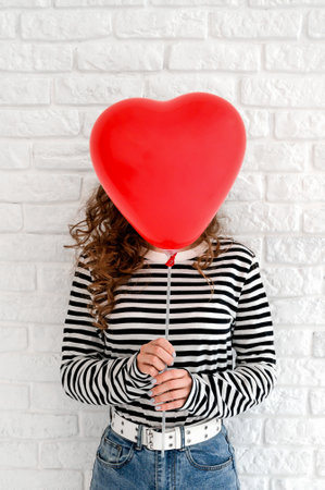 Woman covering her head with big red heart shaped balloon against whote wall backgroundの写真素材