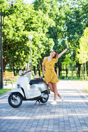 beautiful european girl in a yellow dress with a retro scooter in the morning parkの写真素材