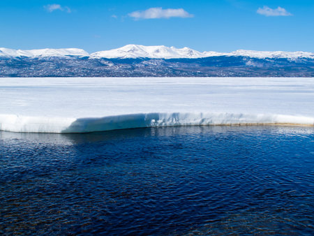 First open water at thawing Lake Laberge, Yukon Territory, Canada, and snow-covered mountains at distant shore of ice-covered frozen lake.の写真素材