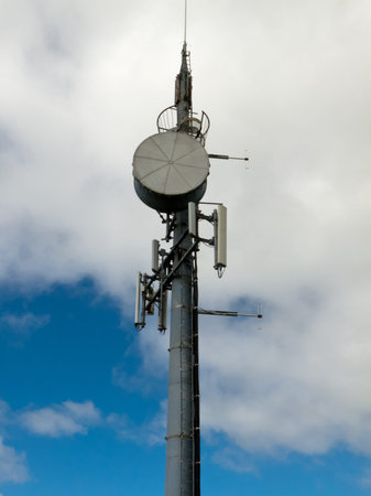 Metal tower with mobile cell phone telecommunications antennas against cloudy blue sky with...