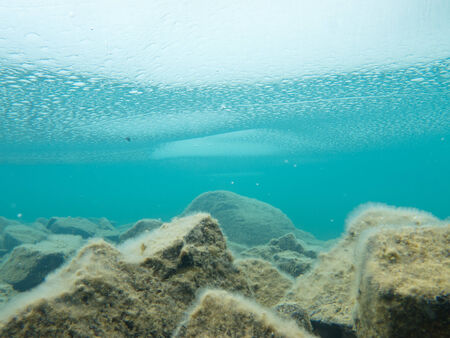 Underwater shot of ice floes floating in clear shallow water with rocky bottom mirrored on water surfaceの写真素材
