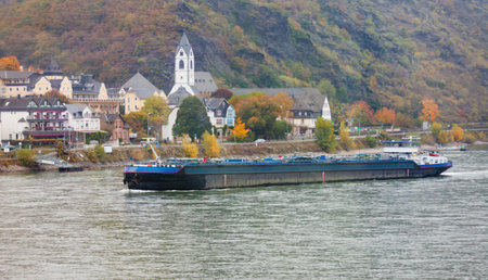 Inland vessel plying waterway river Rhine with small village on background shore, North...