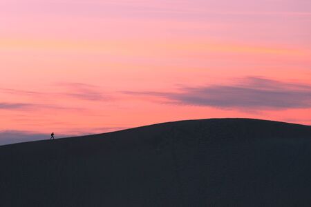 Lone person hiking on crest of huge sand dune, black silhouette against warm colored sunset skyの写真素材