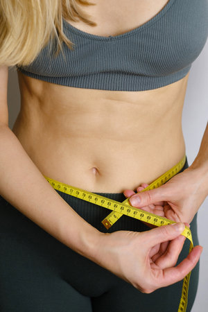 A body centimeter in the hands of girl in tracksuit after a workout or fitness session. Measurement of body volume. Sports waist. Healthy lifestyle, physical activityの写真素材