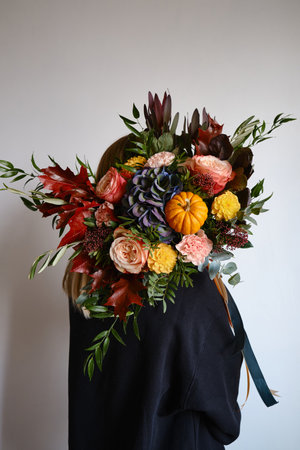Rich bouquet with autumn flowers and a pumpkin in the hands of a girl on a light background. Bright festive bouquet. A holiday gift. Beautiful autumn flowers. Florist girlの写真素材
