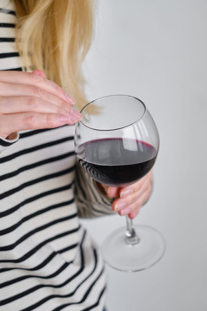 A glass of red wine in the hands of a girl in a striped jacket against the background of a white wall. Wine tasting at the bar. Party, drinks, holidays and celebration conceptの写真素材