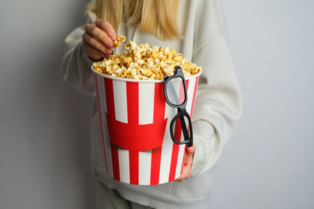 Popcorn paper bucket and 3D glasses in the hands of a young girl preparing to watch a movie. Showtime. Eating delicious unhealthy sweet snacks. Going to cinema for a new filmの写真素材