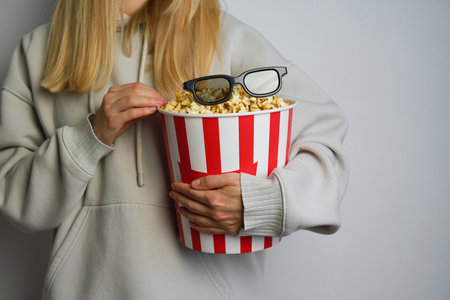 Popcorn paper bucket and 3D glasses in the hands of a young girl preparing to watch a movie. Showtime. Eating delicious unhealthy sweet snacks. Going to cinema for a new filmの写真素材