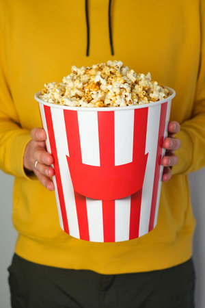 Popcorn paper bucket in the hands of a men preparing to watch a movie. Showtime. Eating delicious unhealthy sweet snacks. Going to cinema for a new film. Rest and entertainmentの写真素材