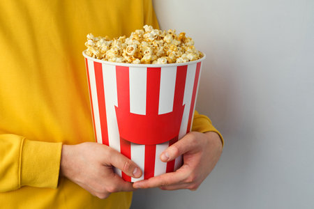 Popcorn paper bucket in the hands of a men preparing to watch a movie. Showtime. Eating delicious unhealthy sweet snacks. Going to cinema for a new film. Rest and entertainmentの写真素材